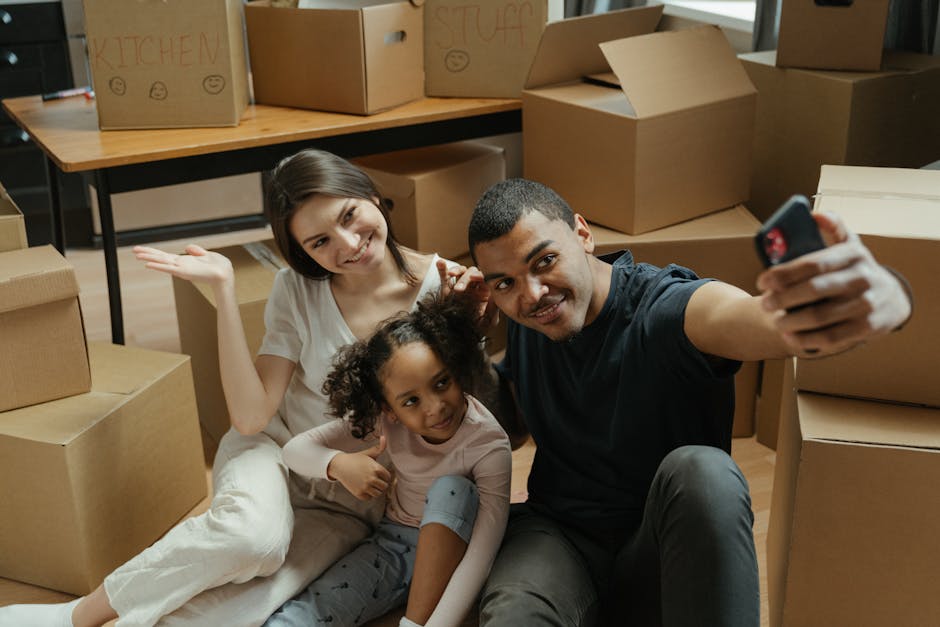 A man and a woman are inside a home surrounded by stacks of brown cardboard moving boxes, some sealed with packing tape. The man, dressed in a white t-shirt and blue jeans, is lifting the woman in a j