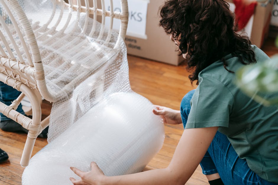 A man with dark curly hair and a beard is standing inside a room during a home relocation, carefully packing fragile glassware into a white cardboard box marked with a red 'FRAGILE' sticker. He is wea