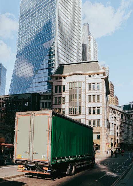 A large moving truck with a green cargo container is parked on a city street, close to the curb, during daylight hours. The truck is positioned near tall modern glass office buildings and historic mul